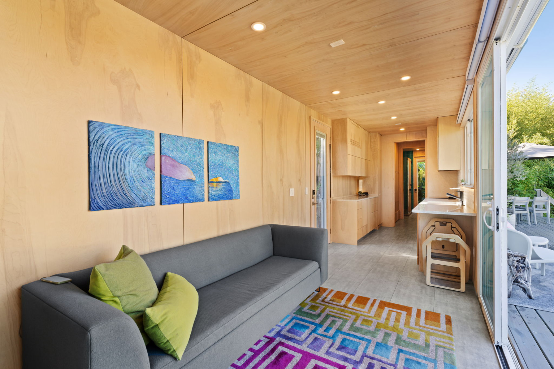 Interior lounge with wood walls, sofa, and kitchen opening onto the deck